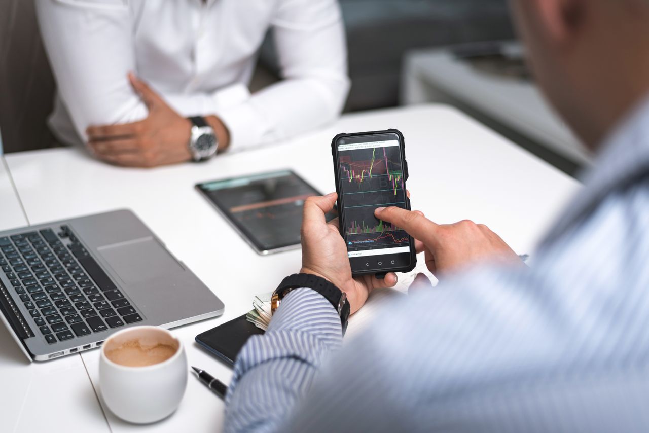 heros-img Two businessmen analyze stock market data using a smartphone and laptop in a modern office setting.